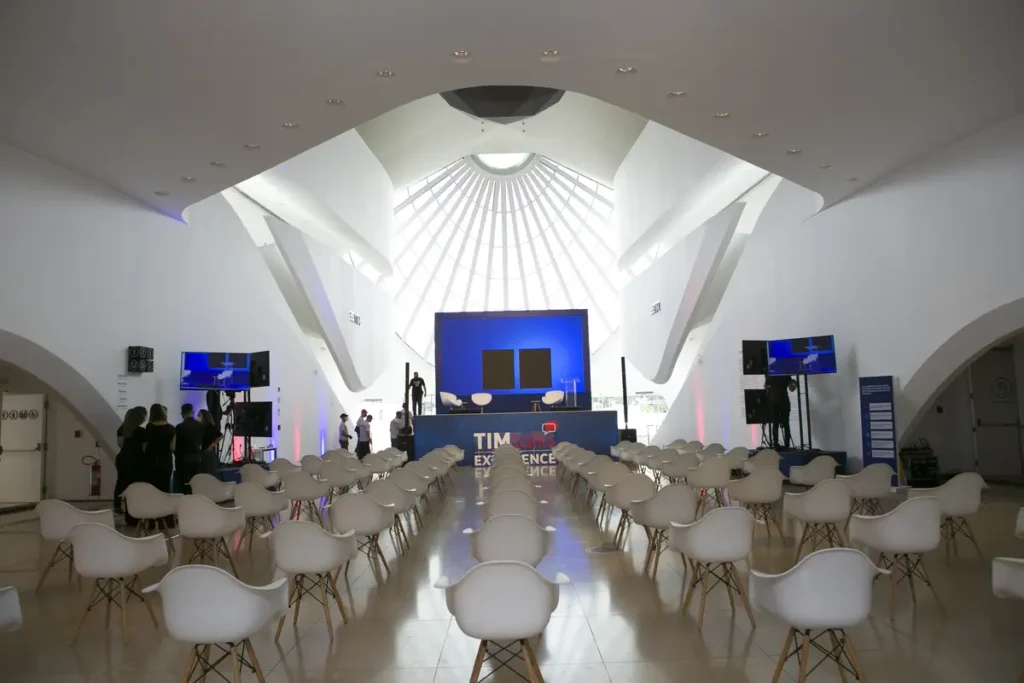 Eventos híbridos: sete desafios e soluções para empresas Sala de evento com cadeiras brancas alinhadas voltadas para palco azul com tela, em ambiente arquitetônico branco e iluminado