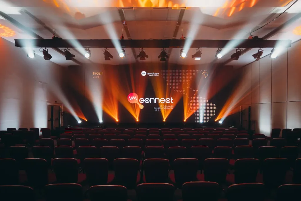 Como Planejar Eventos Corporativos Estratégicos do Zero Auditório com fileiras de cadeiras vazias voltadas para um palco iluminado em tons de laranja e branco com projeção de mapa e logo do evento VM Connect
