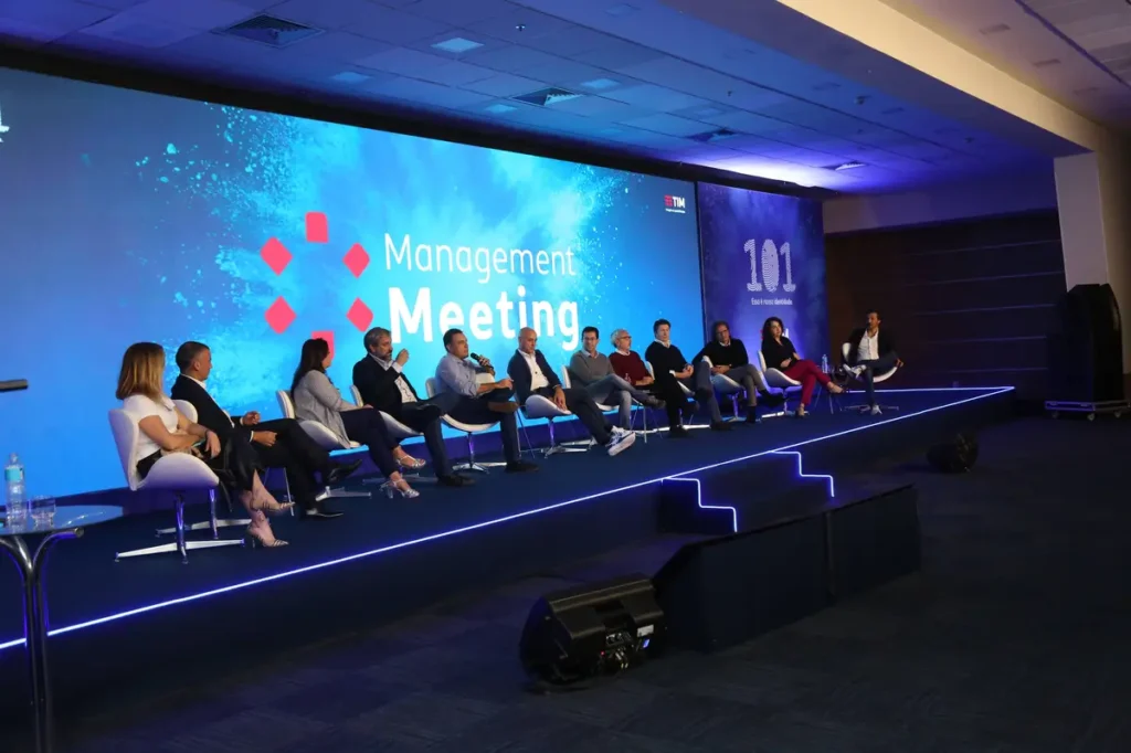 Tipos de eventos corporativos: saiba escolher o ideal para sua empresa Painel de discussão com dez pessoas sentadas em cadeiras em um palco iluminado azul.